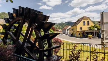 Historisches Wasserrad im Vordergrund mit Blick auf eine Straße und gelbe Gebäude in bergiger Umgebung.