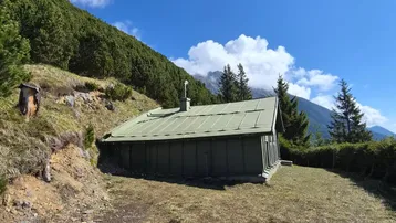 Eine rustikale Hütte mit grünem Satteldach steht auf einer Wiese vor einem bewaldeten Berghang.