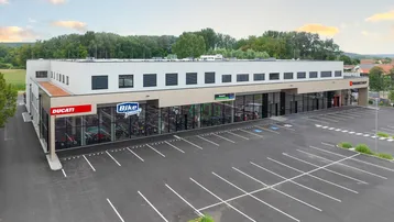 Moderne Gewerbeimmobilie mit großen Schaufenstern und weitläufigem Parkplatz, umgeben von Grünflächen.