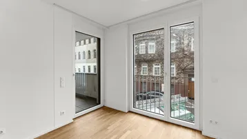 Heller Raum mit Holzboden und großen Fenstern, eines davon als französischer Balkon gestaltet.