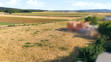 Luftaufnahme eines weiten Feldes mit ländlicher Umgebung und verschwommenen Gebäuden im Hintergrund.