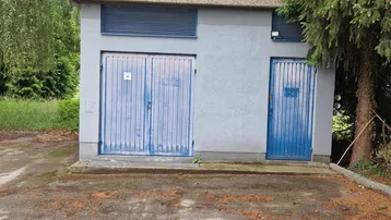 Kleines graues Nebengebäude mit blauen Türen, umgeben von hohen Bäumen und viel Grün.