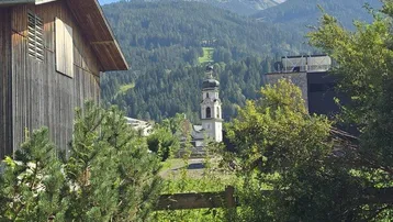 Blick auf eine Kirche und Berge, umgeben von üppigem Grün unter blauem Himmel.