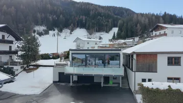 Modernes Gebäude mit großen Glasflächen und Balkon in verschneiter Berglandschaft.