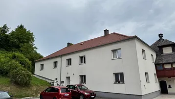 Weiß verputztes Mehrfamilienhaus mit rotem Ziegeldach und Parkplätzen davor.