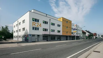 Modernes Gebäude mit weißer und gelber Fassade, Geschäften im Erdgeschoss und Parkplätzen davor.