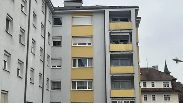Mehrstöckiges Wohngebäude mit grauer Fassade und gelben Balkonen unter bewölktem Himmel.