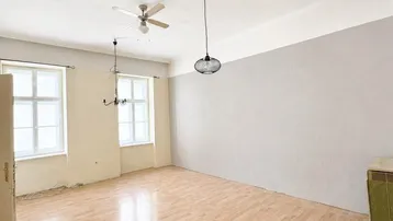 Helles, leeres Zimmer mit Parkettboden, zwei Fenstern und Deckenventilator, bereit zur Renovierung.