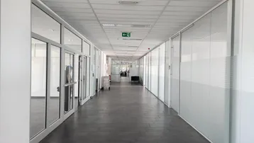 Langer, heller Büroflur mit Glaswänden und dunklem Fliesenboden, der zu verschiedenen Büros führt.