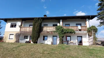 Gut gepflegtes Haus mit mehreren Balkonen und einem grünen Hang im Vordergrund unter blauem Himmel.