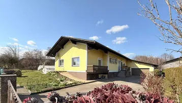 Gelbes Einfamilienhaus mit Satteldach, umgeben von einem Garten und einer Einfahrt an einem sonnigen Tag.