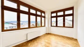Großzügiger Raum mit vielen Fenstern, Fischgrätparkett und weitem Ausblick, ideal als Wohnzimmer.