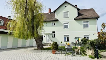 Grünes Mehrfamilienhaus mit gepflastertem Vorplatz, Garagen und einem großen Baum.
