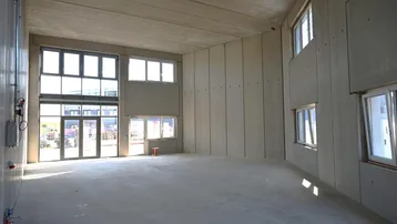 Großer, heller Innenraum mit hohen Decken und vielen Fenstern, noch im Rohbauzustand.