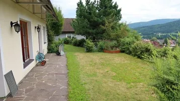 Gepflasterte Terrasse neben dem Haus mit Rasenfläche und weitem Blick in die bergige Landschaft.