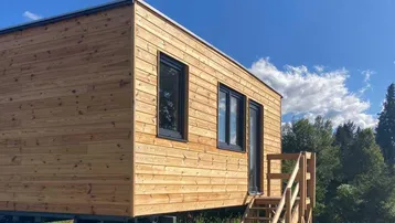 Modernes Tiny House mit Holzfassade und schwarzem Flachdach unter blauem Himmel.