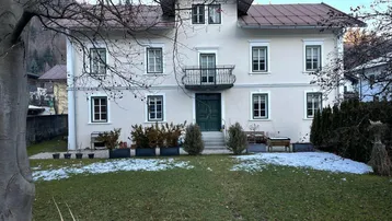 Klassisches Mehrfamilienhaus mit Balkon und Vorgarten in einer bergigen Umgebung.