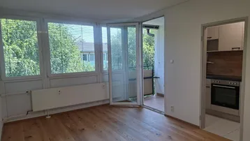 Heller Raum mit Holzboden, großen Fenstern und Zugang zu einem kleinen Balkon, daneben eine Kochnische.