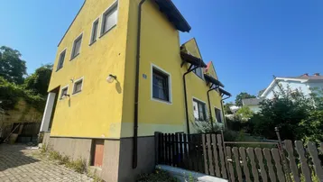 Gelbes Einfamilienhaus mit gepflastertem Zugang und Holzzaun unter blauem Himmel.