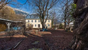 Historisches Gebäude mit gelber Fassade und blauem Dach, umgeben von Bäumen und einem Gartenbereich mit Bergblick.