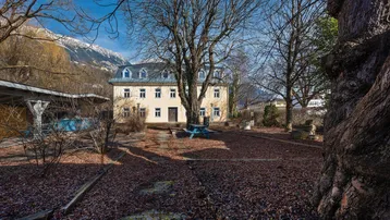 Historisches Gebäude mit Mansarddach, umgeben von kahlen Bäumen und Blick auf schneebedeckte Berge.
