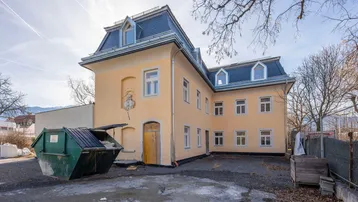 Gelbes Gebäude mit Mansarddach und einem Müllcontainer auf dem Vorplatz unter blauem Himmel.