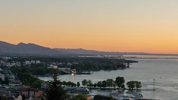 Panoramablick auf eine Stadt am Seeufer bei Sonnenuntergang, mit Bergen im Hintergrund und ruhigem Wasser.