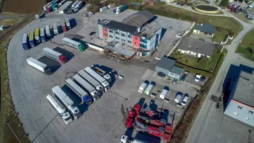 Luftaufnahme eines Gewerbeparks mit Hauptgebäude, Lagerhallen und großem LKW-Parkplatz in ländlicher Umgebung.
