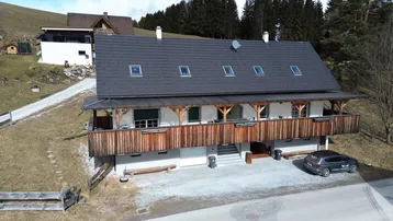 Haus mit Holzfassade und langem Balkon in alpiner Umgebung mit Parkplatz.