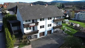 Mehrfamilienhaus mit mehreren Balkonen und Garagen in ländlicher Umgebung, umgeben von Grün.