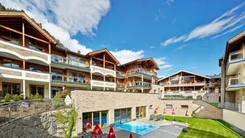 Mehrere Gebäude im alpinen Stil mit Außenpool und gepflegter Grünfläche unter blauem Himmel.