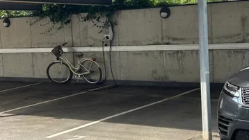 Überdachter Parkplatz mit Ladestation für Elektrofahrzeuge und Fahrrad, umgeben von Betonwänden und Bäumen.