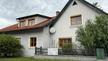 Seitenansicht des Hauses mit Satteldach, Holzfenstern und einem Verkaufsschild im Vorgarten.