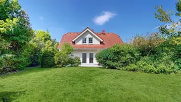 Vorderansicht des Hauses mit gepflegtem Rasen und üppiger grüner Bepflanzung unter blauem Himmel.