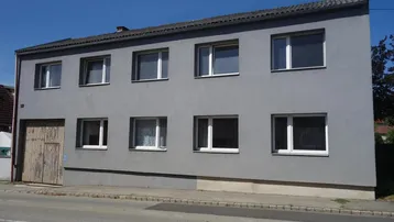 Ansicht der grauen Fassade eines zweistöckigen Gebäudes mit mehreren Fenstern und einer großen Holztür.