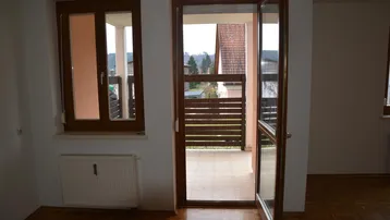 Heller Innenraum mit Parkettboden, Fenster und Zugang zu einem Außenbereich mit Blick ins Grüne.