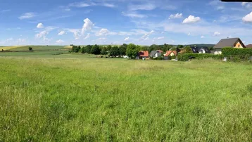 Weitläufige grüne Landschaft mit Feldern und Häusern am Horizont unter blauem Himmel.
