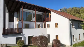 Modernes Haus mit großen Glasfronten und Balkonen, umgeben von Grünflächen unter blauem Himmel.