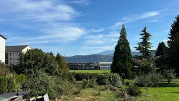 Blick über ein grünes Grundstück mit Bäumen und Bergen im Hintergrund unter blauem Himmel.