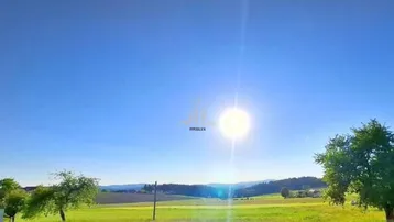 Sonnige Landschaft mit weiten Feldern, Bäumen und sanften Hügeln in der Umgebung.