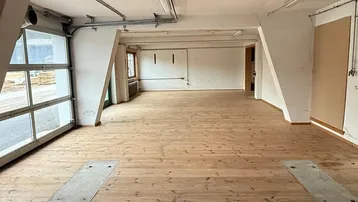 Großer, leerer Raum mit Holzdielenboden und einem Garagentor, das viel Tageslicht hereinlässt.