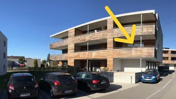 Modernes Mehrfamilienhaus mit Holzfassade und Balkonen, ein Pfeil zeigt auf eine Wohnung.