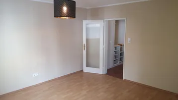 Helles Zimmer mit Parkettboden und Zugang zu einem weiteren Raum mit Regalen.