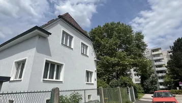 Helles Einfamilienhaus mit gepflegtem Vorgarten und Blick auf die Umgebung unter blauem Himmel.