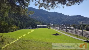 Weitläufige grüne Wiese mit bewaldeten Bergen im Hintergrund und Blick auf die umliegende Landschaft.
