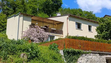 Hausfassade mit Balkon und üppiger Vegetation in sonniger Hanglage.