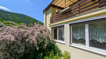 Nahaufnahme der Hausfassade mit Balkon und blühenden Sträuchern vor grüner Bergkulisse.