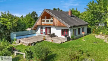 Freistehendes Haus mit Holzterrasse, Pool und umgebender Grünfläche unter blauem Himmel.