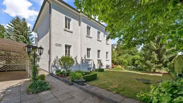 Weißes Wohnhaus mit gepflegtem Garten, großer Rasenfläche, altem Baumbestand und gepflasterter Zufahrt.
