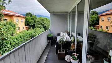 Möblierter Balkon mit Holzboden und Blick ins Grüne sowie auf umliegende Gebäude.